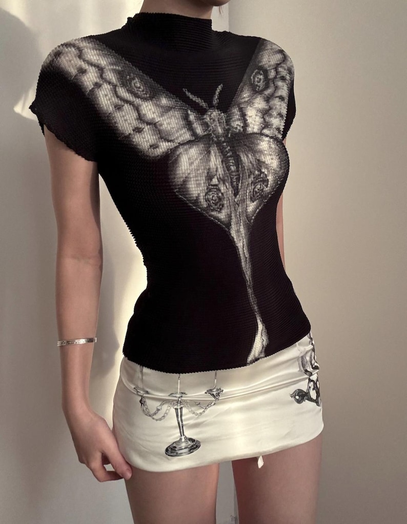 May include: A black short-sleeved top with a high neck and a large white moth graphic across the chest, paired with a cream-colored mini skirt featuring a black and white illustration of a candelabra and other decorative elements on the fabric.