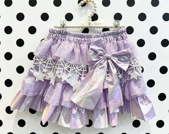 May include: A lavender tiered skirt featuring layers of shimmering tulle and metallic fabric. The skirt is adorned with white beaded fringe, embroidered star appliques, and a large, prominent metallic lavender bow on the front right side.