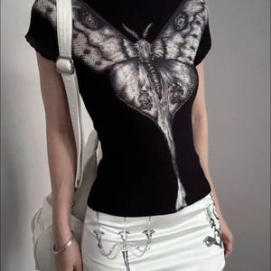 May include: A black short-sleeved mock neck top featuring a large, detailed grey moth graphic on the front. Paired with a white mini skirt that displays a printed illustration of a silver candelabra and a white shoulder bag with a silver buckle.