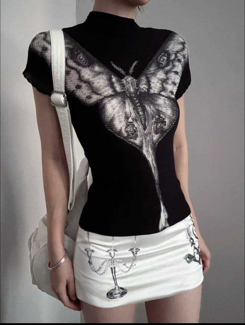 May include: A black short-sleeved mock neck top featuring a large, detailed grey moth graphic on the front. Paired with a white mini skirt that displays a printed illustration of a silver candelabra and a white shoulder bag with a silver buckle.