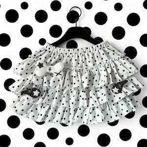 Può includere: Una gonna bianca a pois neri caratterizzata da tre livelli di balze. La gonna è decorata con un'applicazione in peluche bianco a forma di osso sul livello superiore e due applicazioni nere a forma di stella con pois bianchi sui livelli inferiori.