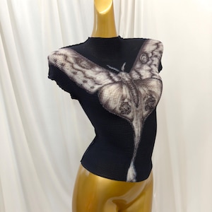 May include: A black short-sleeved top with a high neckline and a textured, pleated fabric surface. The front features a large, detailed graphic print of a moth in shades of white, gray, and black, extending from the chest down to the waist.