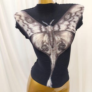 May include: A black, high-neck, cap-sleeve top with a textured, pleated fabric surface. The front features a large, detailed graphic print of a moth in shades of white, gray, and black, with its wings spanning across the chest and shoulders.