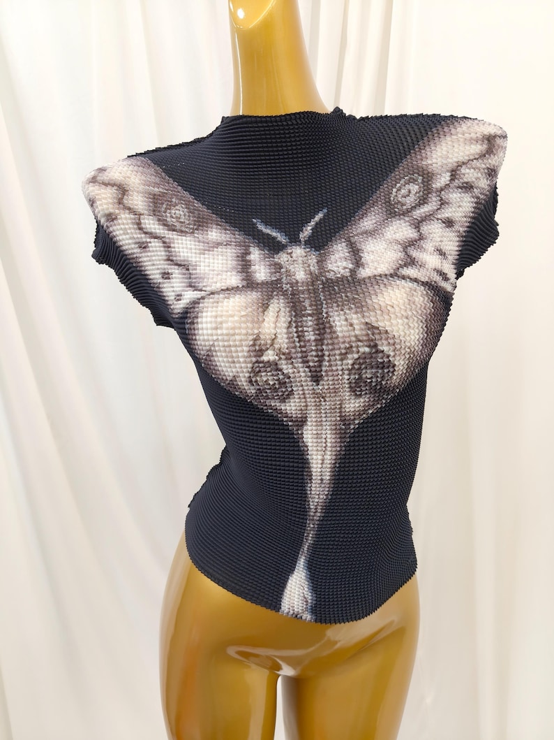 May include: A black, high-neck, cap-sleeve top with a textured, pleated fabric surface. The front features a large, detailed graphic print of a moth in shades of white, gray, and black, with its wings spanning across the chest and shoulders.