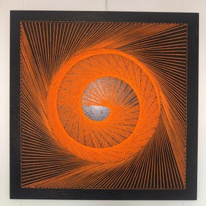 Op de afbeelding: Een vierkant draadkunstwerk met een zwart houten frame en ondergrond. Feloranje draad is rond kleine spijkers gewikkeld om een geometrisch spiraalpatroon te creëren dat uitstraalt vanuit een centrale cirkelvormige negatieve ruimte.