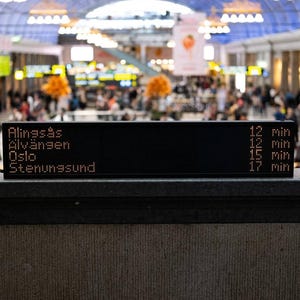 Può includere: Un tabellone digitale nero per le partenze dei treni che mostra le destinazioni Alingsås, Älvängen, Oslo e Stenungsund con i relativi tempi di arrivo in minuti, su uno sfondo sfocato di un affollato terminal pubblico.
