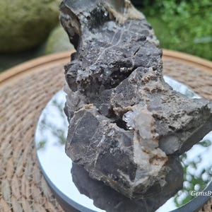 Op de afbeelding: Een ruw, donkergrijs mineraal exemplaar met een ruwe, onregelmatige textuur en grillige randen. Kleine, doorschijnende, kristallijne formaties zijn ingebed in de spleten van de steen. De tekst GemsByTamar is zichtbaar in de rechterbenedenhoek.
