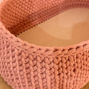 Könnte beinhalten: Ein runder Aufbewahrungskorb mit dicken, altrosa gehäkelten Kordelwänden und einem glatten, hellen Holzboden. Die Seiten sind in einem dichten, geflochtenen Maschenmuster gearbeitet, das einen strukturierten und stabilen Rand an der Oberseite bildet.