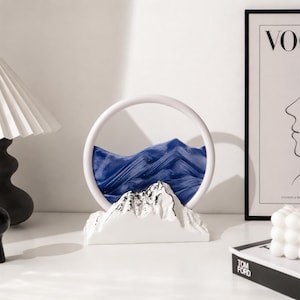 May include: A circular sand art display featuring a white frame and a white mountain-shaped base. Inside the glass circle, blue sand forms layered, wave-like patterns that resemble a mountain range against a dark blue background.