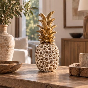Könnte beinhalten: Eine dekorative Ananas-Figur mit einer goldfarbenen Spitze aus spitzen Blättern und einem Körper, der mit kleinen, weißen, blumenförmigen Muscheln bedeckt ist, die in einem dichten, sich wiederholenden Muster auf der Oberfläche angeordnet sind.