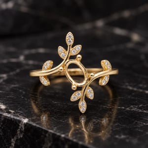 Puede incluir: Un anillo de oro con un engaste ovalado central para una piedra, flanqueado por dos ramas simétricas en forma de hoja. Cada hoja está acentuada con pequeñas piedras redondas transparentes incrustadas en la superficie del metal.