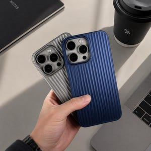 Könnte beinhalten: Zwei Smartphone-Hüllen mit vertikaler Rillenstruktur. Eine Hülle ist dunkelblau, die andere silberfarben. Beide Hüllen verfügen über einen quadratischen Kameraausschnitt mit drei kreisförmigen Linsen und einem Blitzlicht.
