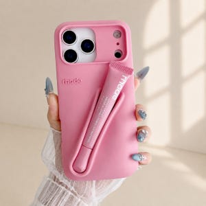 Puede incluir: Una funda de silicona para smartphone en color rosa mate con un bolsillo exterior moldeado diseñado para sujetar un tubo a juego de bálsamo labial rhode peptide lip tint. La palabra rhode está grabada en relieve y tiene aberturas para la cámara.