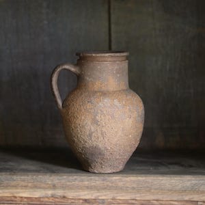May include: A rustic, brown ceramic pitcher with a textured, weathered surface. The vessel features a rounded body, a short neck with a defined rim, and a single curved handle attached to the side. The finish appears matte and uneven in tone.