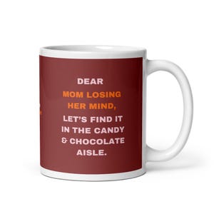 Op de afbeelding: Een witte keramische mok met een donkerrode buitenkant voorzien van de tekst in lichtroze en oranje: DEAR MOM LOSING HER MIND, LET'S FIND IT IN THE CANDY & CHOCOLATE AISLE. De mok heeft een gebogen handvat aan de rechterkant en een gladde afwerking.
