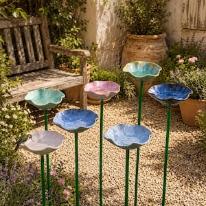 May include: A collection of six ceramic bird baths on thin green metal stakes, featuring scalloped, bowl-shaped tops in various shades of blue, turquoise, and white. Each basin is mounted on a slender pole, creating a tiered garden display.