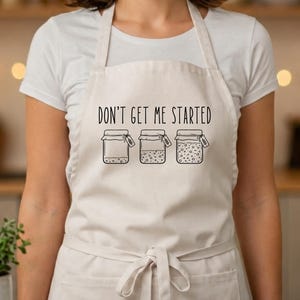 May include: A beige apron featuring a black graphic print of three glass jars in varying stages of fermentation. Above the jars, the text reads DON'T GET ME STARTED in a simple, clean font. The apron includes a waist tie and a front pocket.