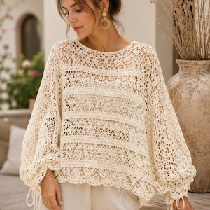 May include: A cream-colored crochet top featuring a loose, airy knit pattern with horizontal bands of intricate lace detailing. The garment has long, voluminous bishop sleeves that gather at the wrists with adjustable drawstring ties and a round neckline.