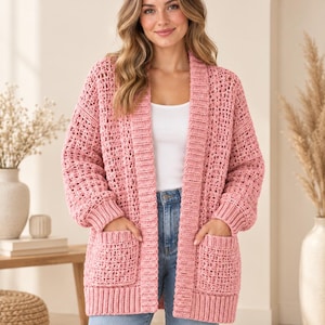 Könnte beinhalten: Eine altrosa Strickjacke mit offener Vorderseite, grober Häkelstruktur, langen Ärmeln mit gerippten Bündchen, einem breiten gerippten Kragen und zwei großen aufgesetzten Vordertaschen. Das Kleidungsstück wird über einem weißen Top und einer hellblauen Jeans getragen.