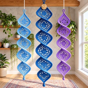 Peut inclure: Trois décorations suspendues en spirale composées de motifs carrés au crochet dans des tons bleu clair, bleu moyen et violet. Chaque pièce verticale présente une série de motifs floraux reliés, suspendus par des cordons blancs avec une étiquette en bois.
