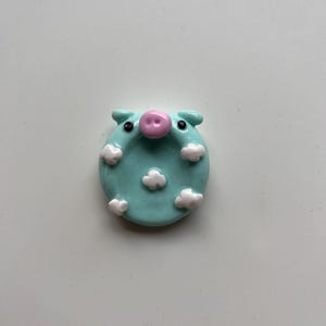 May include: A circular light blue ceramic trinket dish shaped like a pig face. The dish features a raised pink snout, two small black eyes, and white cloud-like shapes scattered across the surface. The piece has a smooth, glossy finish.