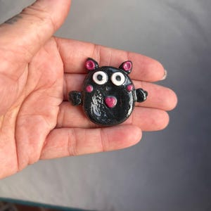 Könnte beinhalten: Eine kleine, schwarze, abgerundete Kreaturenfigur mit glänzender Oberfläche. Sie hat zwei große weiße Augen mit schwarzen Pupillen, kleine rosa Ohren, rosa Wangenflecken und ein herzförmiges rosa Detail auf der Brust. Die Figur hat zwei kleine Arme.