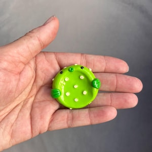 May include: A small, round, lime green ceramic dish shaped like a pufferfish. The dish features a smooth, glossy finish, two dark eyes, a small protruding mouth, two side fins, and several small, raised white dots across its surface.