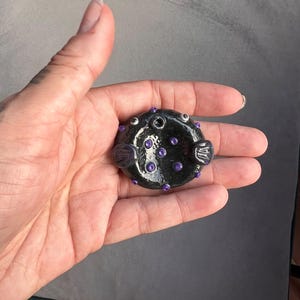May include: A small, round, black ceramic object shaped like a pufferfish with a glossy finish. It features two small white eyes, two side fins, and several small, raised purple circular accents embedded across its surface.