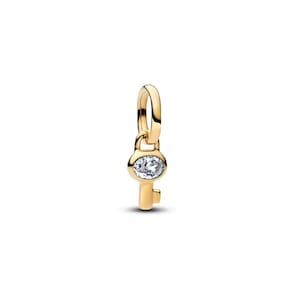 May include: A gold-toned metal charm shaped like a key, featuring a small oval-shaped clear gemstone set into the head of the key. The charm includes a smooth, polished finish and a circular loop at the top for attachment to a chain or bracelet.