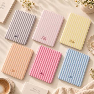 Pode incluir: Seis cadernos retangulares com riscas verticais em cinzento, rosa claro, amarelo, laranja, rosa choque e azul claro. Cada caderno apresenta um nome ou iniciais personalizados impressos no centro: AM, Ella, Mia, Leo, Ava e XO.
