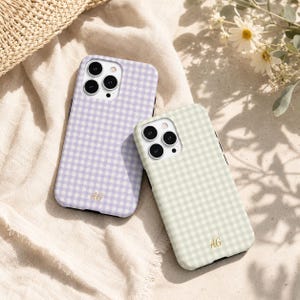 May include: Two smartphone cases featuring a gingham check pattern in light purple and light green. Each case has a triple camera cutout and the gold-coloured initials AG printed in a script font near the bottom edge of the back surface.