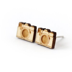 Op de afbeelding: Een paar houten camera-oorbellen. De oorbellen zijn gemaakt van lichtbruin hout en hebben een donkerbruine omtrek. Het cameradesign is eenvoudig en minimalistisch.