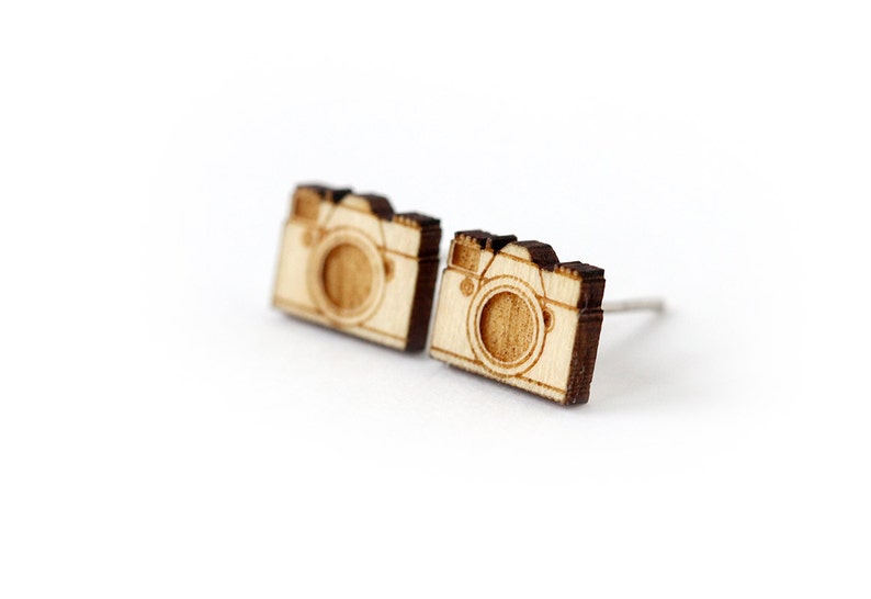 Op de afbeelding: Een paar houten camera-oorbellen. De oorbellen zijn gemaakt van lichtbruin hout en hebben een donkerbruine omtrek. Het cameradesign is eenvoudig en minimalistisch.