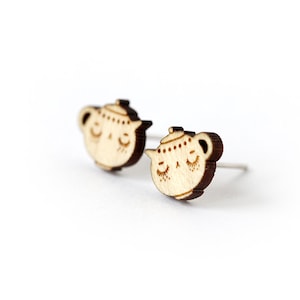 May include: A pair of wooden stud earrings. Each earring is shaped like a teapot with a smiling face and closed eyes. The earrings are light brown with silver posts, set against a white background.