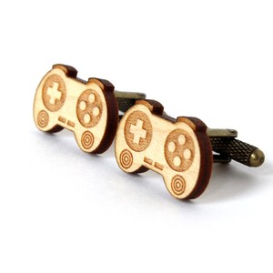May include: A pair of wooden cufflinks shaped like a video game controller. The cufflinks are brown and have a light wood grain. The controller has a plus sign button and four circle buttons.