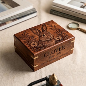 May include: A rectangular wooden box with a dark brown finish featuring a laser-engraved rabbit surrounded by wildflowers on the lid. The front side displays the text CLOVER 2018-2026 Forever hopping in our hearts with light-coloured corner accents.