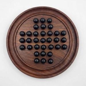 May include: A circular dark brown wooden board game featuring a cross-shaped grid of holes. The board contains thirty-two smooth, spherical black game pieces arranged in a pattern, leaving the center hole empty for a solitaire peg game.