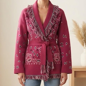 Könnte beinhalten: Eine magentafarbene Strickjacke mit dunklem Paisle mit dunklem Paisle mit dunklem Paisleymuster, einem Schalkragen mit Fransenbesatz und einem passenden Bindegürtel in der Taille. Das langärmelige Kleidungsstück hat einen geraden Saum mit zusätzlichen Fransen am unteren Rand.