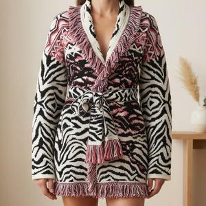 May include: A long-sleeved knit cardigan featuring a black, white, and pink zebra-inspired pattern. The garment includes a shawl collar, a matching tie-waist belt, and decorative fringe trim along the collar, hem, and belt ends.