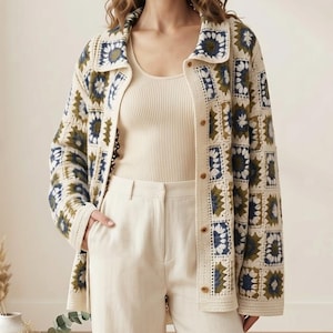 May include: A long-sleeved cardigan featuring a multi-colored granny square crochet pattern in shades of cream, dark blue, and olive green. The garment has a collared neckline, a button-down front, and a ribbed hem, worn over a cream ribbed tank top.