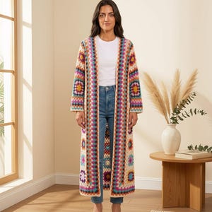 May include: A long, open-front cardigan featuring a colorful granny square crochet pattern with cream, pink, orange, blue, and purple yarn. The garment is worn over a plain white t-shirt and blue denim jeans, extending down to the lower calf area.