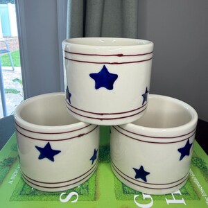 May include: Three cylindrical ceramic crocks with a cream-colored glaze. Each piece features a hand-painted dark blue star pattern around the center and two thin, horizontal dark red stripes encircling the top and bottom rims of the vessels.