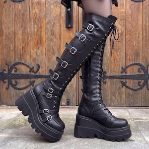 May include: Black knee-high platform boots featuring a thick lug sole, a lace-up front with silver-toned eyelets, and multiple horizontal straps across the shaft secured with silver metal buckles. The boots have a smooth finish and a wedge heel.