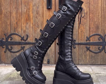 May include: Black knee-high platform boots featuring a thick lug sole, a lace-up front with silver-toned eyelets, and multiple horizontal straps across the shaft secured with silver metal buckles. The boots have a smooth finish and a wedge heel.