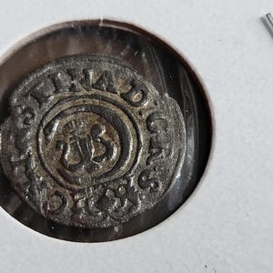 Puede incluir: Una moneda circular de metal plateado desgastado con un símbolo central en relieve que se asemeja a una flor de lis o corona estilizada. La moneda tiene la inscripción STINA D.G. AS en el borde exterior y está en un soporte protector.