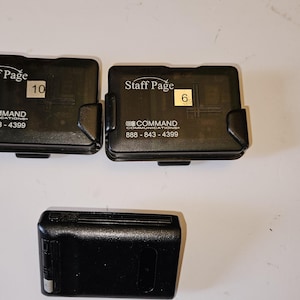 Puede incluir: Tres buscapersonas electrónicos rectangulares de color negro. Dos dispositivos muestran el texto Staff Page, Command Communications y un número de teléfono, con etiquetas blancas numeradas 10 y 6. El tercer dispositivo se sitúa debajo.