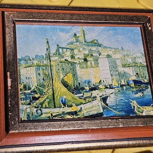 May include: A framed oil painting depicting a harbor scene with sailboats, figures on a boat, and a hillside city in the background. The frame is dark brown with a reddish inner border and shows signs of wear on the edges. The painting uses blue, yellow, and white tones to capture the water, sky, and buildings.