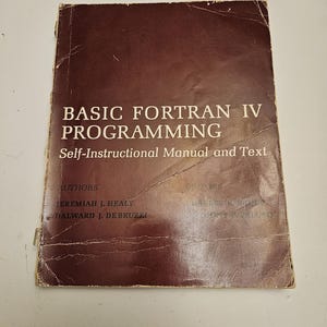Può includere: Un libro rettangolare con copertina color marrone scuro intitolato BASIC FORTRAN IV PROGRAMMING Self-Instructional Manual and Text. La copertina elenca gli autori Jeremiah J. Healy e Dalward J. Debruzzi, mostrando segni di usura con pieghe e bordi rovinati.