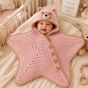 Könnte beinhalten: Eine hellrosa, sternförmige Häkeldecke für Babys mit einer Kapuze, die ein Bärengesicht und Ohren aufweist. Die Decke hat einen beigefarbenen Rand und wird mit fünf braunen Holzknöpfen entlang der diagonalen Kante verschlossen.