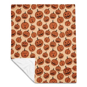 May include: A soft throw blanket featuring a repeating pattern of orange jack-o'-lanterns with black hand-drawn facial expressions on a light beige background. The blanket is folded at one corner to reveal a solid white underside.